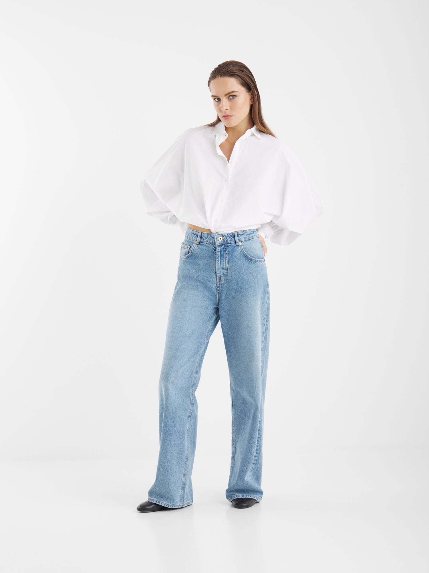 Eine Frau steht vor einem weißen Hintergrund und trägt ein weißes Hemd mit Knopfleiste und eine hellblaue Jeans mit hoher Taille und weiten Beinen aus Denim. Sie hat glattes braunes Haar, einen neutralen Gesichtsausdruck und die Hände in den Taschen ihrer Jeans.