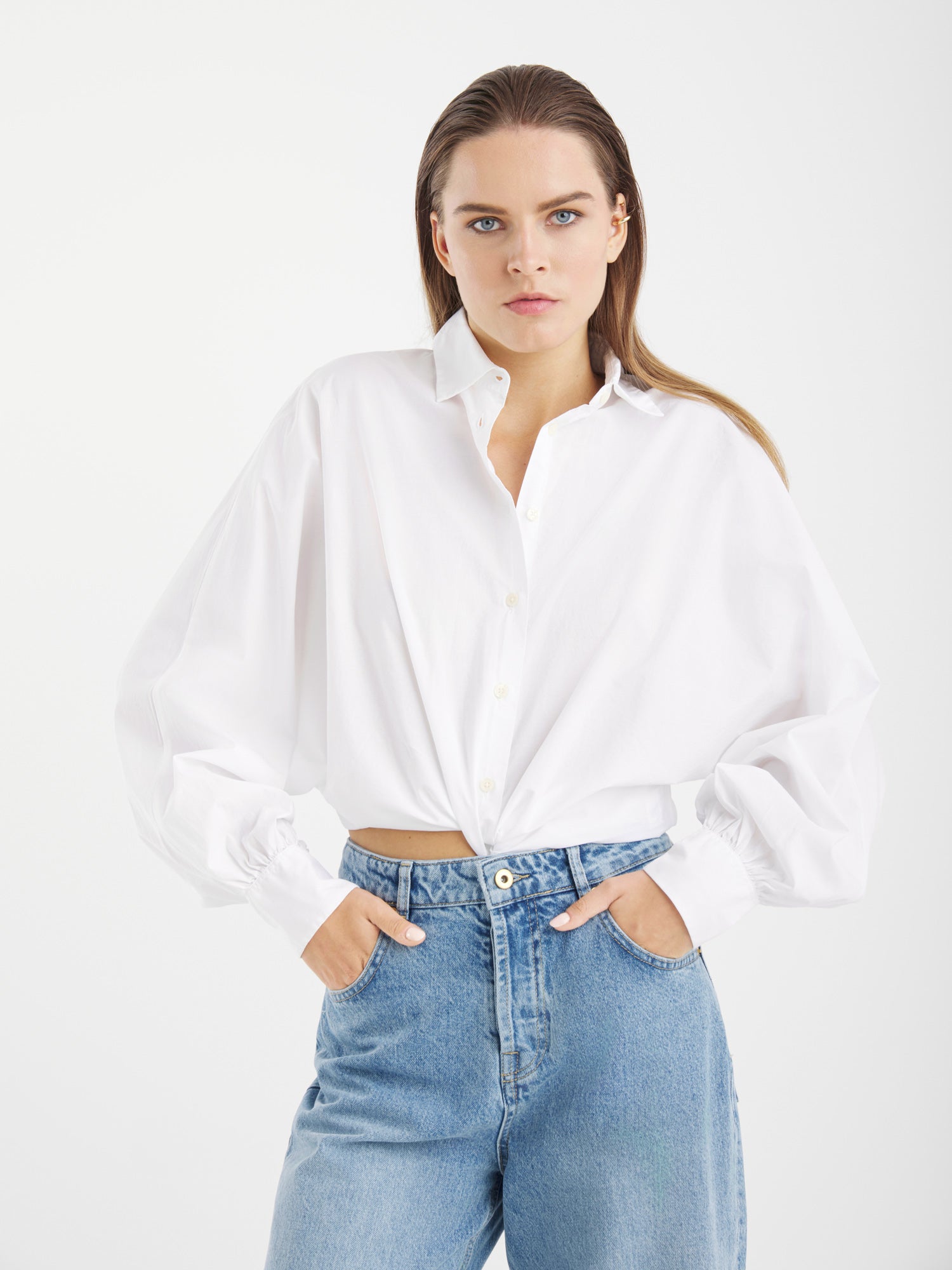 Eine Person mit glattem, hellbraunem Haar trägt die Bluse Antonia Slim White - ein locker sitzendes weißes Hemd aus 100 % Baumwolle, das in eine hochgekrempelte Blue Jeans gesteckt wird. Sie steht vor einem schlichten weißen Hintergrund. Hergestellt in Portugal.