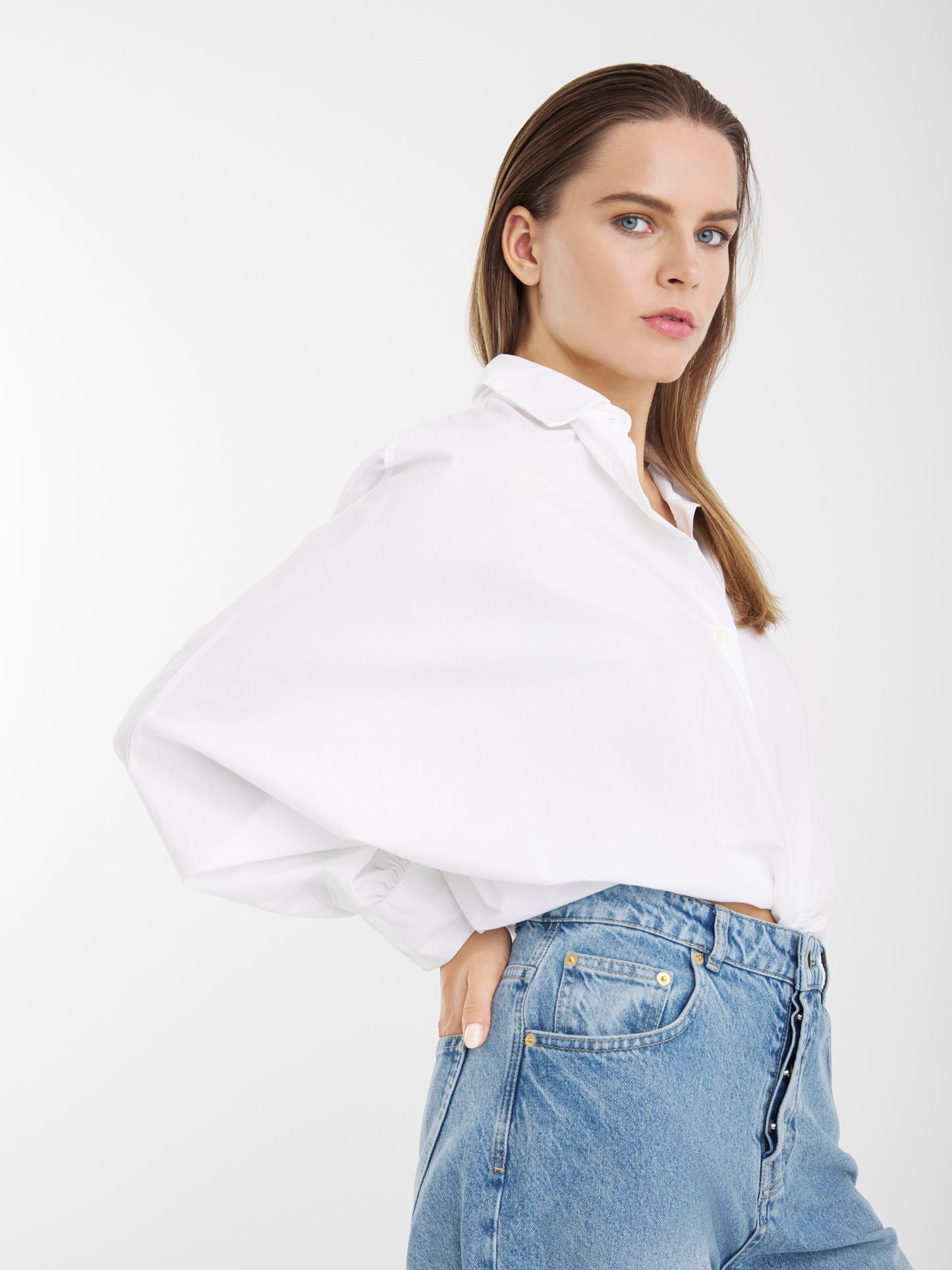 Eine Frau mit glattem braunem Haar trägt die Bluse Antonia Slim White, die in Portugal aus 100 % Baumwolle hergestellt wird, und blaue Jeans mit hoher Taille. Sie posiert mit einer Hand an der Hüfte vor einem schlichten weißen Hintergrund.
