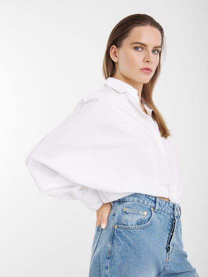 Eine Frau mit glattem braunem Haar trägt die Bluse Antonia Slim White, die in Portugal aus 100 % Baumwolle hergestellt wird, und blaue Jeans mit hoher Taille. Sie posiert mit einer Hand an der Hüfte vor einem schlichten weißen Hintergrund.