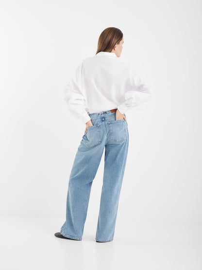 Eine Person steht mit dem Gesicht zur Wand, trägt ein weißes, langärmeliges Hemd und eine hochtaillierte hellblaue Jeans Denim mit weiten Beinen, eine Hand in der Gesäßtasche, vor einem schlichten weißen Hintergrund.