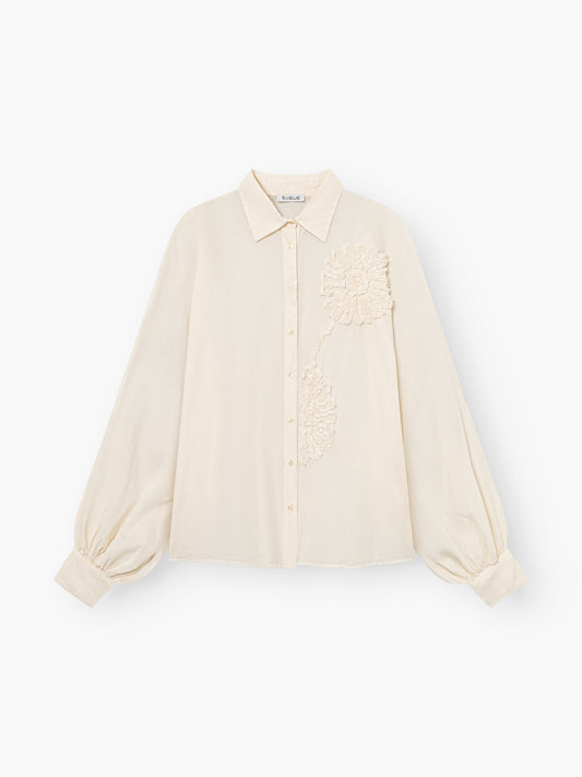 Die Bluse Antonia Crafted Panna ist eine cremefarbene Bluse mit langen, lockeren Ärmeln, die an den Manschetten gerafft sind, einem klassischen Kragen, einer Knopfleiste und einer strukturierten Blumenapplikation auf der oberen linken Seite.