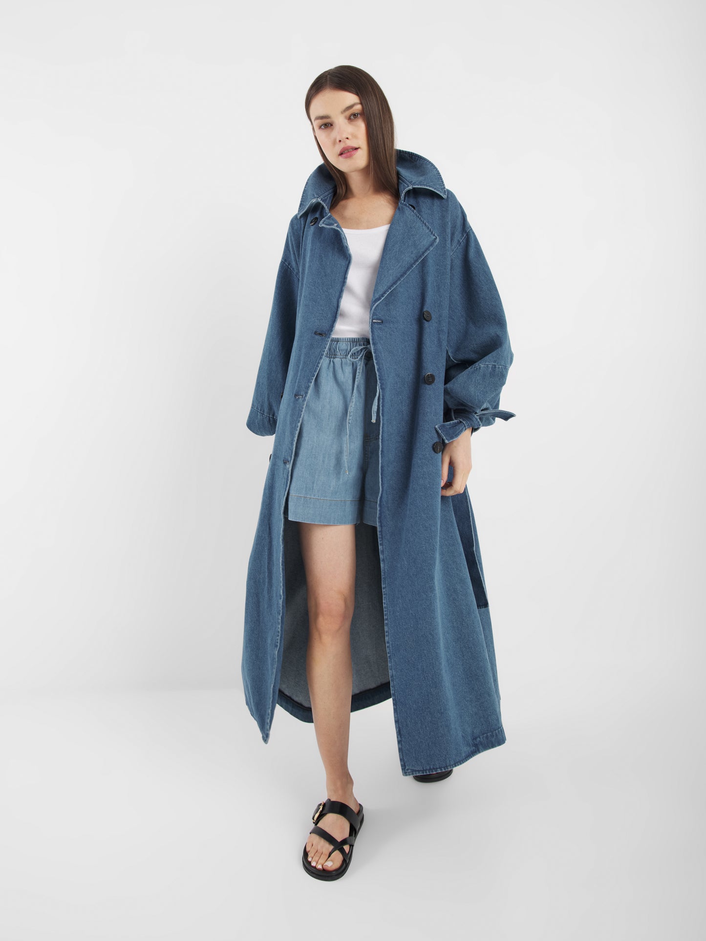 Eine Frau mit glattem braunem Haar und neutralem Gesichtsausdruck trägt den Antonia Denim Trench mit Statement-Ärmeln über einem weißen Oberteil und Jeans-Shorts, gepaart mit schwarzen Sandalen. Sie steht vor einem schlichten weißen Hintergrund. One Size Passform.