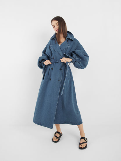 Eine Person steht vor einem unifarbenen weißen Hintergrund und trägt den Antonia Denim Trench mit Statement-Ärmeln, doppelreihigen Knöpfen, einem Gürtel in der Taille, entspannter Passform in Einheitsgröße und schwarzen Sandalen.