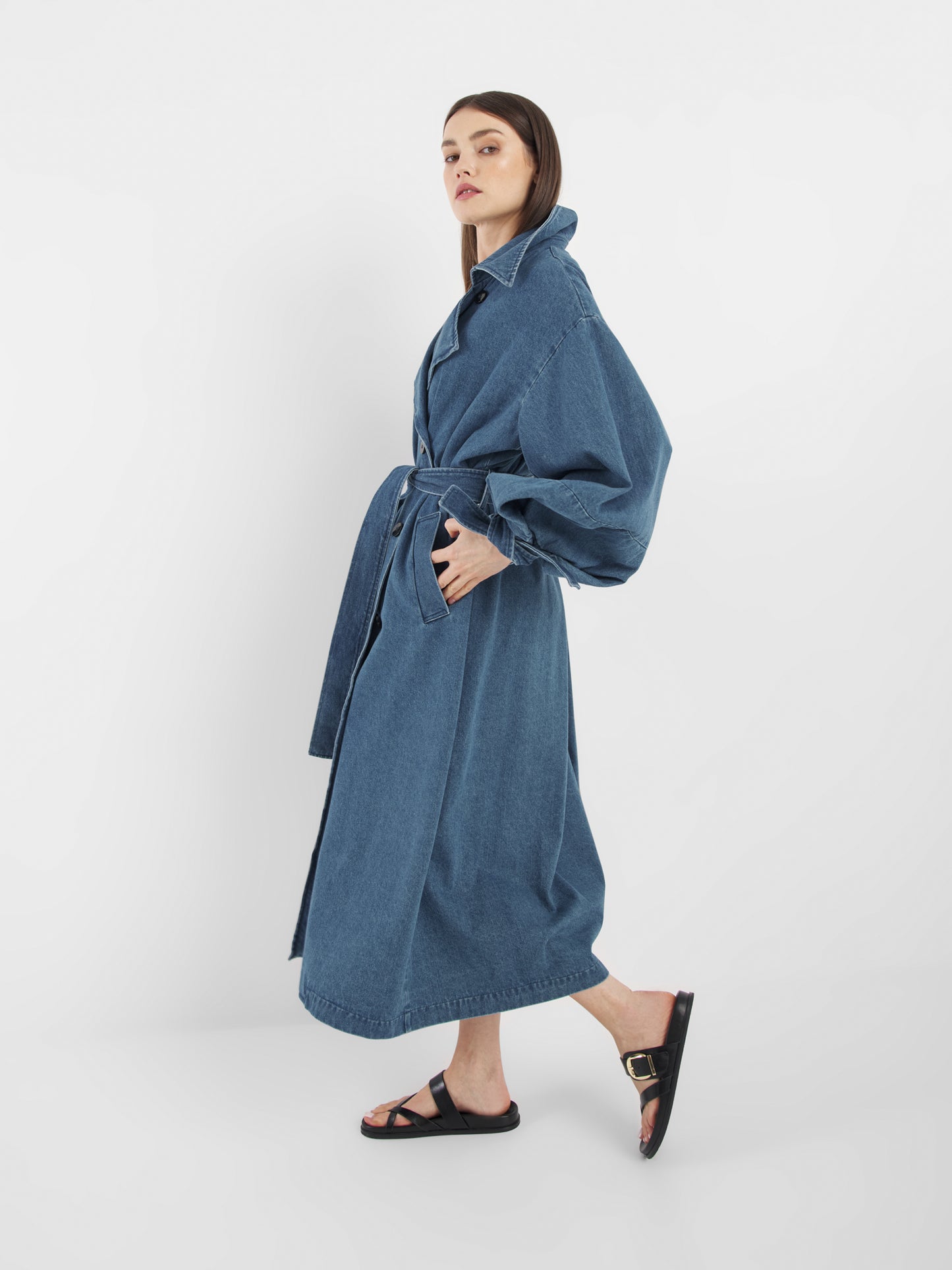 Eine Frau mit dem Antonia Denim Trench (Einheitsgröße) und schwarzen Slide-Sandalen steht im Profil vor einem schlichten weißen Hintergrund.