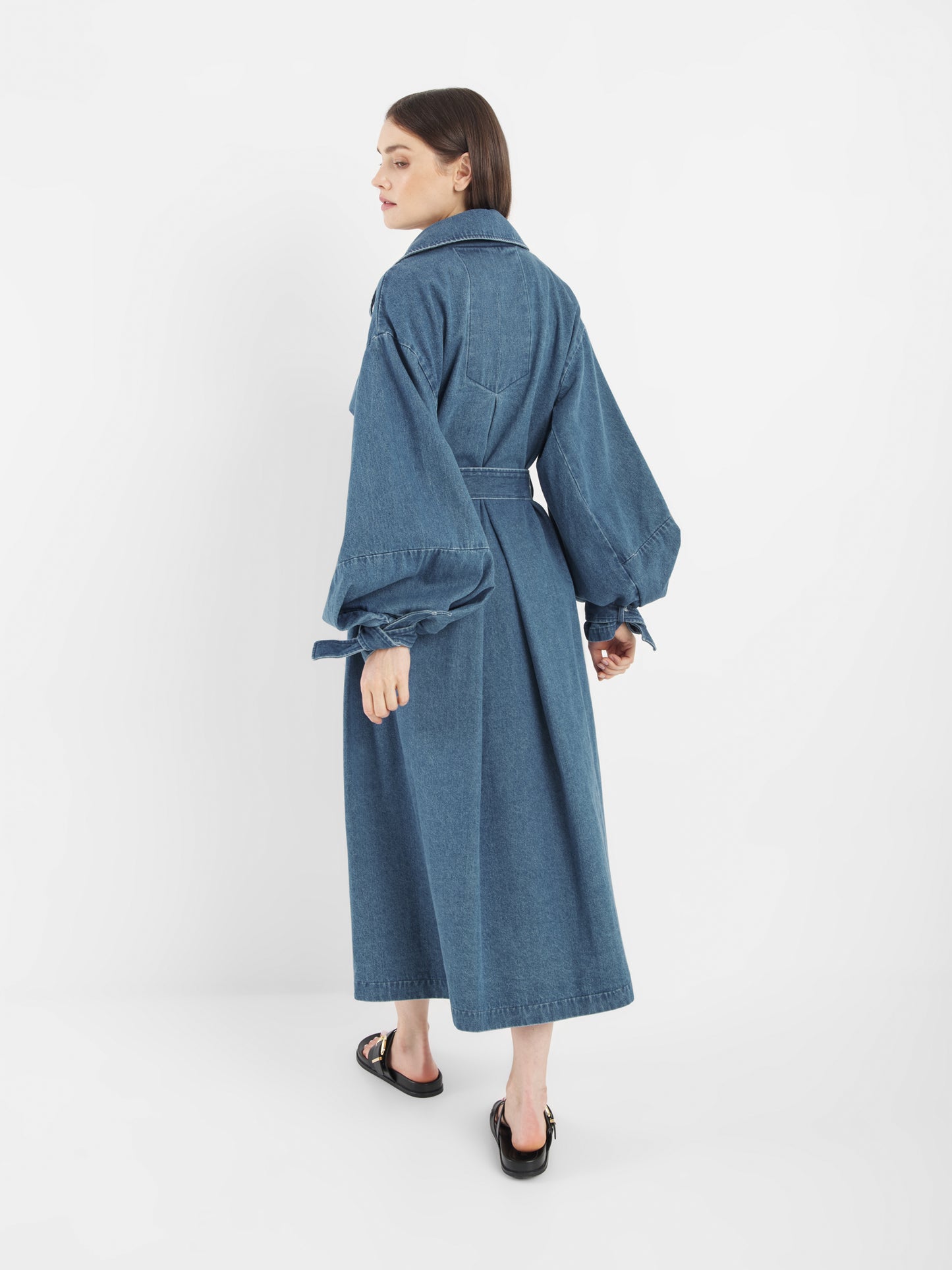Eine Frau steht mit dem Antonia Denim Trench mit Statement-Ärmeln da. Sie hat glattes, schulterlanges Haar und schwarze, offene Schuhe vor einem schlichten weißen Hintergrund.