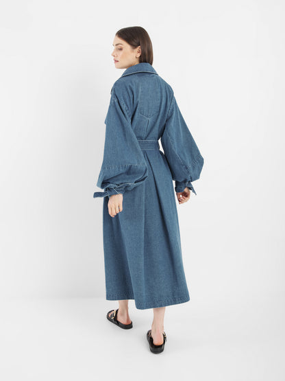 Eine Frau steht mit dem Antonia Denim Trench mit Statement-Ärmeln da. Sie hat glattes, schulterlanges Haar und schwarze, offene Schuhe vor einem schlichten weißen Hintergrund.