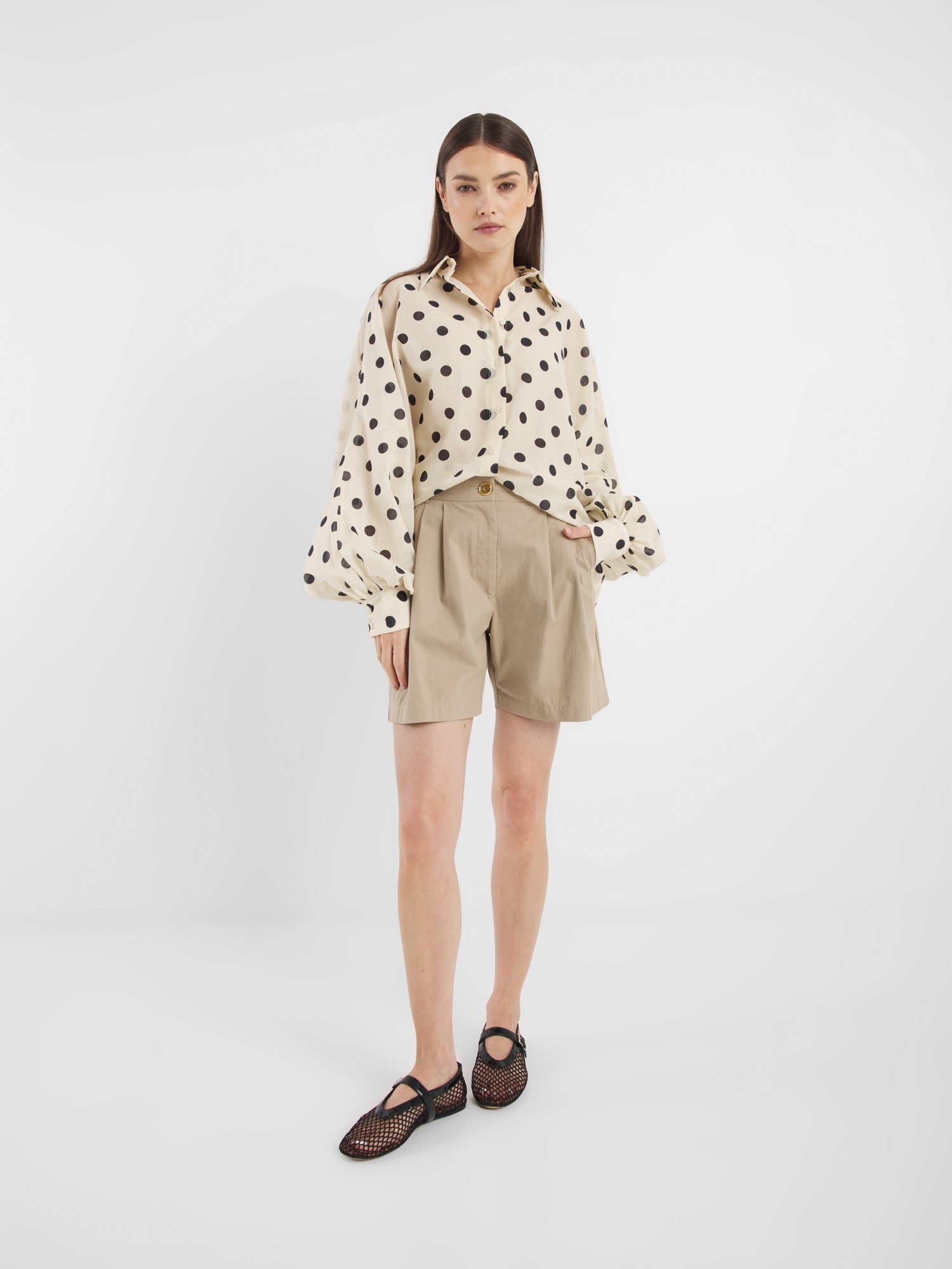Eine Frau mit glattem braunem Haar und neutralem Gesichtsausdruck steht vor einem weißen Hintergrund und trägt beigefarbene, hoch taillierte Shorts Safari, eine cremefarbene Bluse mit schwarzen Punkten und schwarze Netzschuhe.