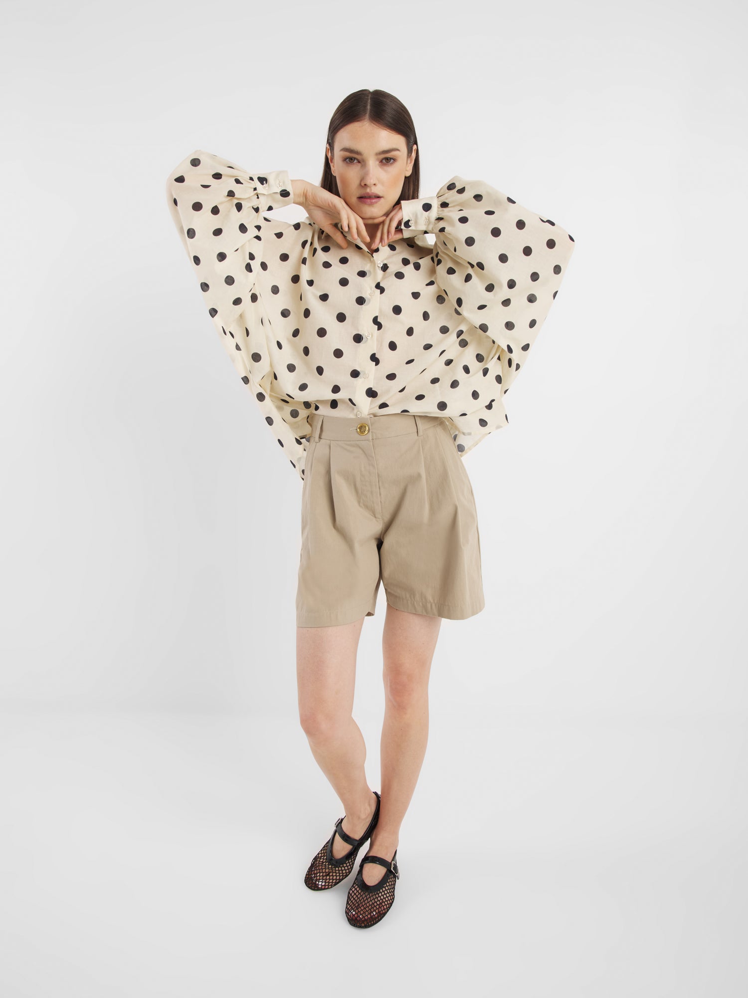 Eine Frau posiert vor einem schlichten weißen Hintergrund und trägt die Bluse Antonia Dots Ecru Black, eine übergroße Baumwoll-Voile-Bluse mit schwarzen Tupfen, gepaart mit beigen Shorts mit hoher Taille und schwarzen Netzschuhen. Ihre Arme sind in der Nähe ihres Kinns erhoben, während sie in die Kamera schaut.