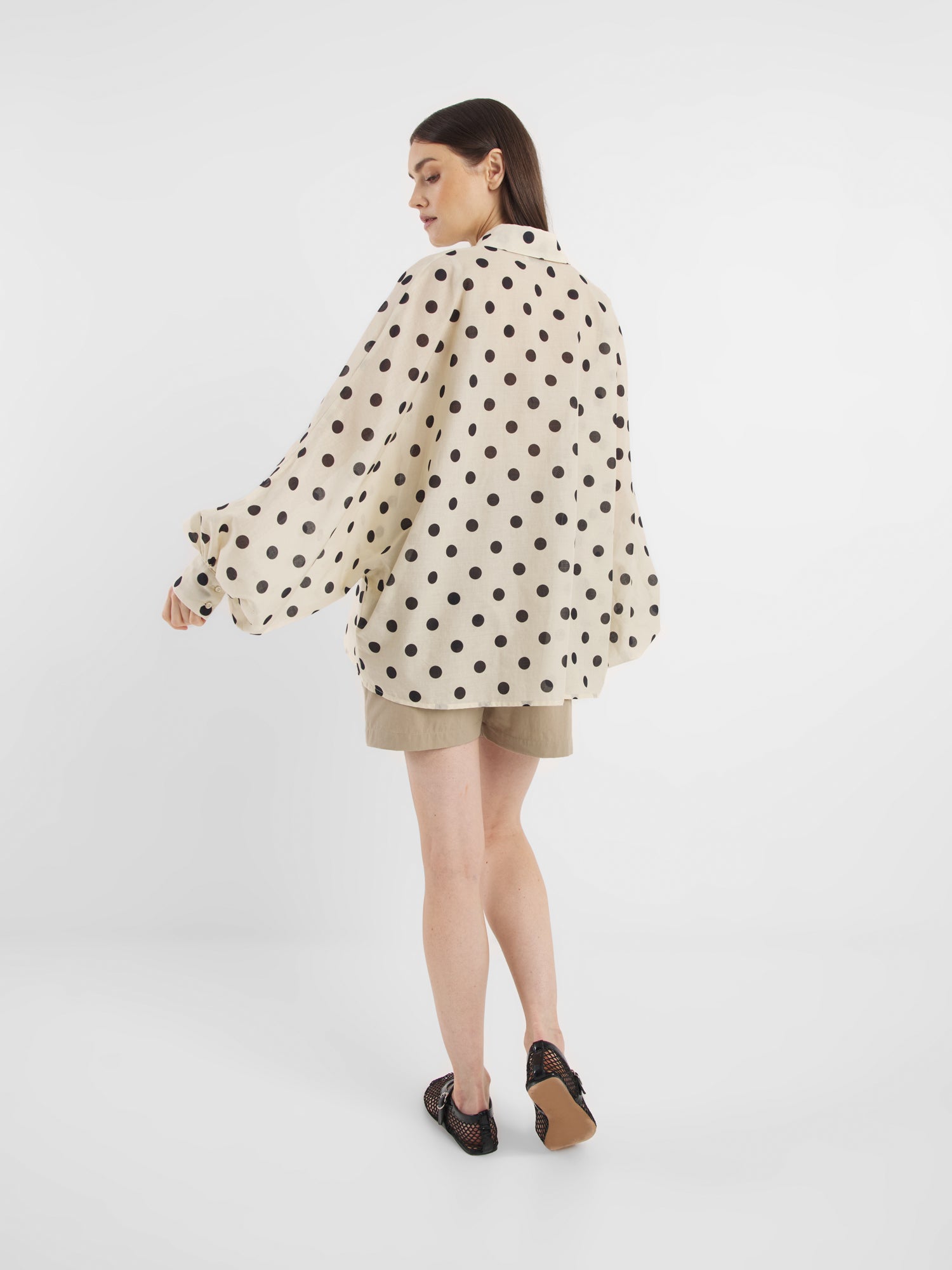 Eine Frau steht in der Bluse Antonia Dots Ecru Black - einer übergroßen Baumwollbluse mit schwarzen Tupfen - mit beigen Shorts und schwarzen Sandalen für einen schicken Sommerlook Damen vor einem schlichten weißen Hintergrund.