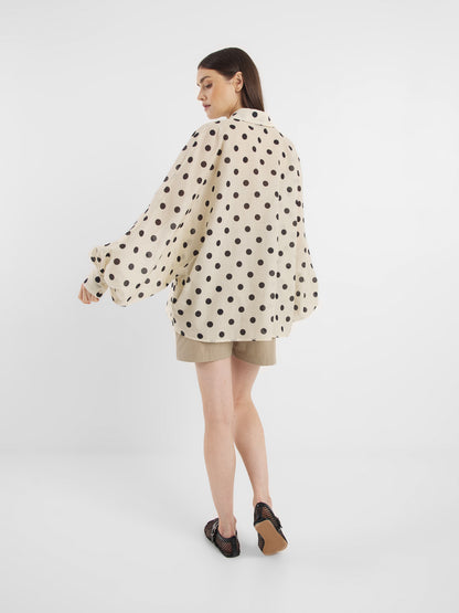 Eine Frau steht in der Bluse Antonia Dots Ecru Black - einer übergroßen Baumwollbluse mit schwarzen Tupfen - mit beigen Shorts und schwarzen Sandalen für einen schicken Sommerlook Damen vor einem schlichten weißen Hintergrund.