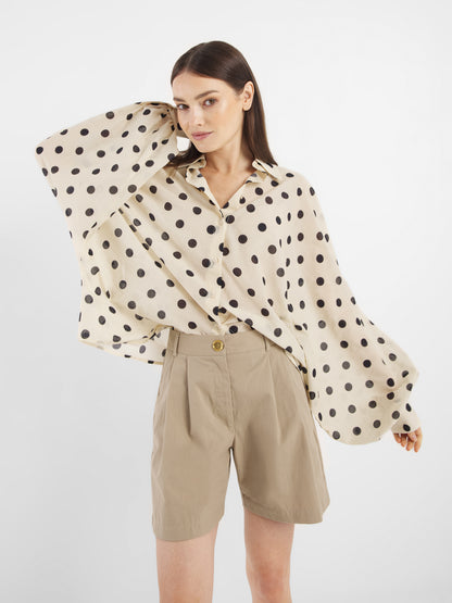 Eine Frau modelliert die übergroße Bluse Antonia Dots Ecru Black mit weiten, fließenden Ärmeln und braunen High-Waisted-Shorts und posiert vor einem schlichten weißen Hintergrund für einen schicken Sommerlook Damen.