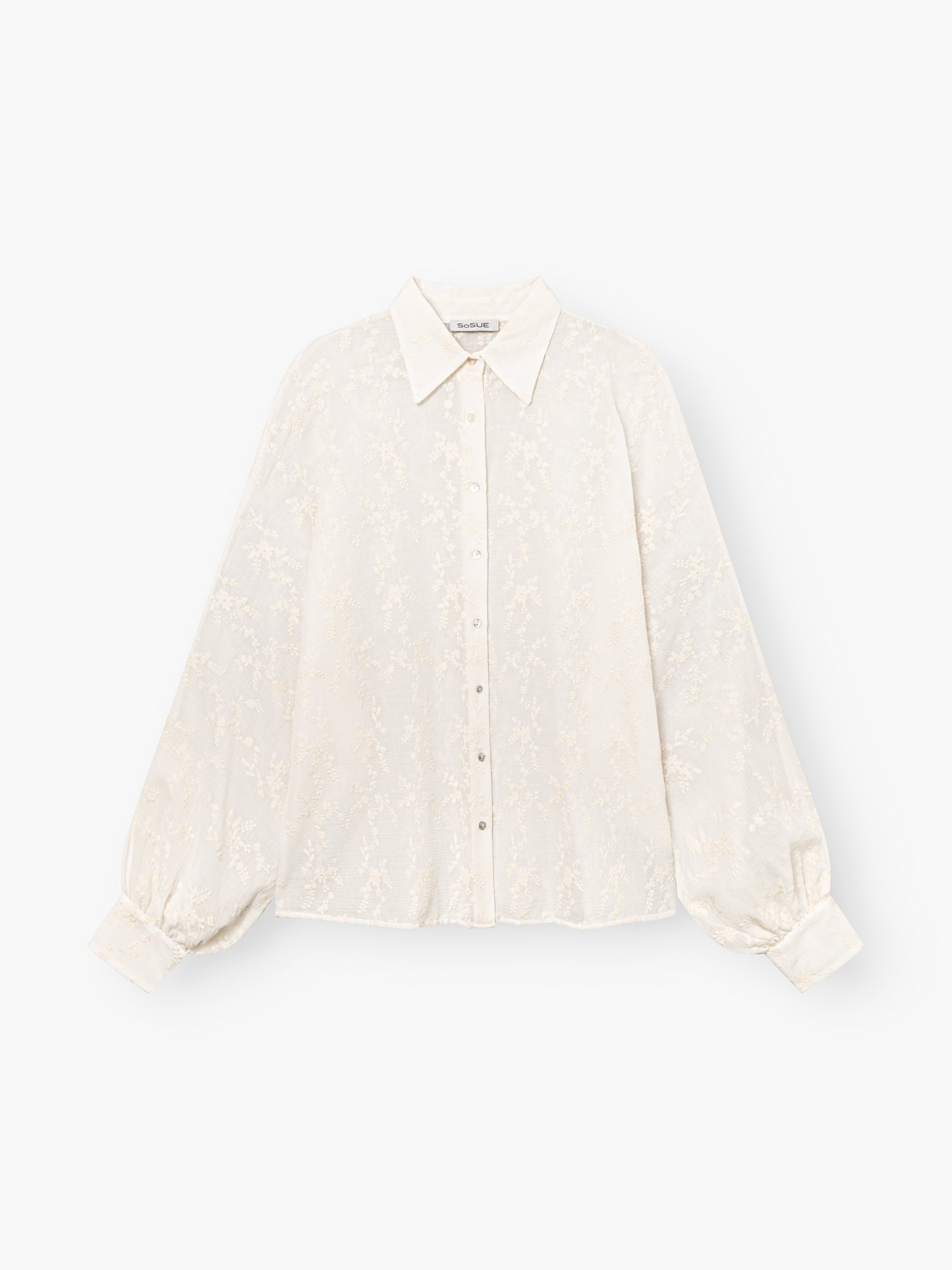 Die Bluse Antonia Embrodery Creme ist eine cremefarbene, langärmelige Bluse mit dezentem Blumenmuster, spitzem Kragen, geknöpfter Vorderseite und breiten Manschetten, abgebildet auf weißem Grund.