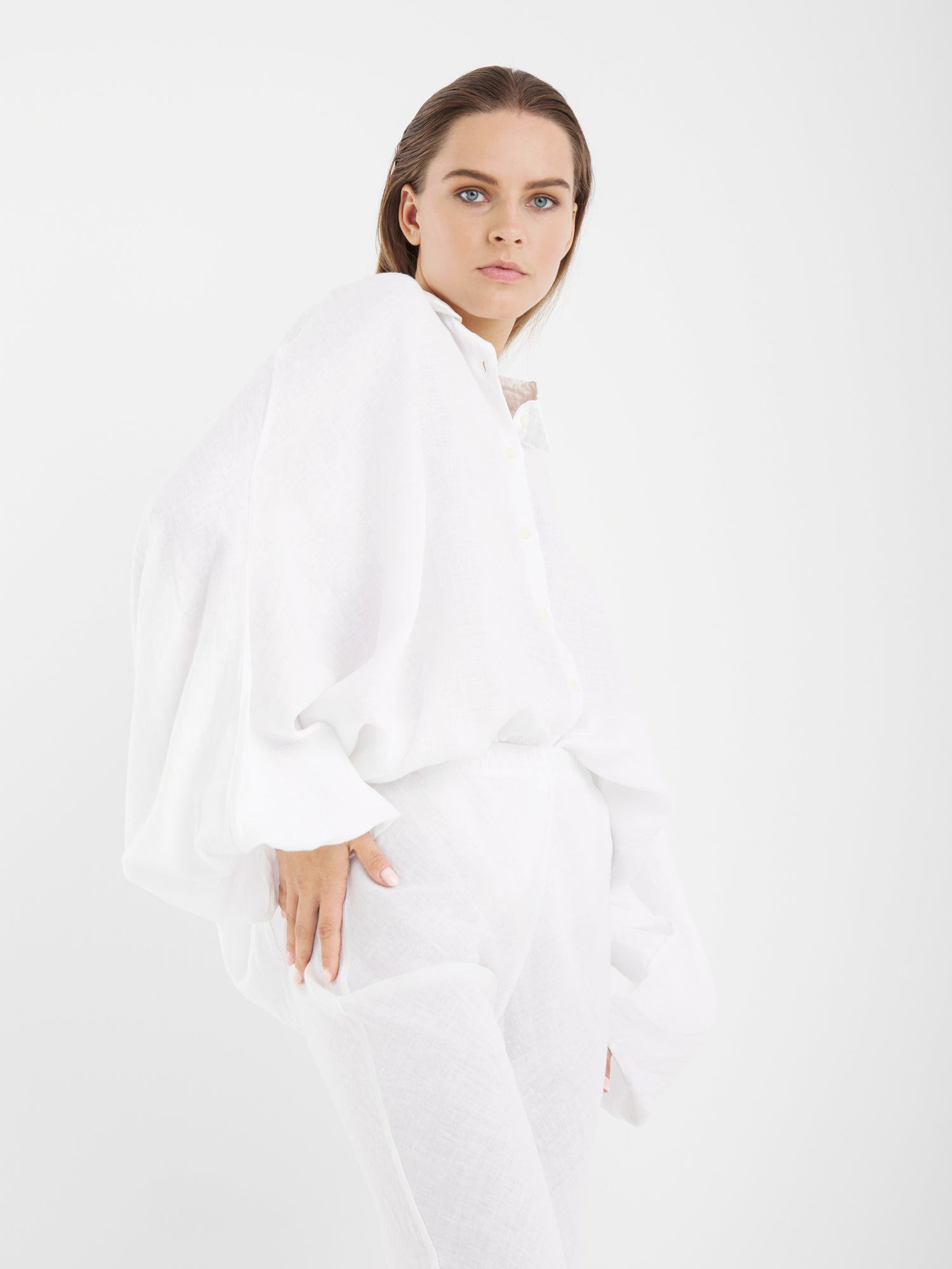 Eine Frau mit heller Haut und glattem braunem Haar posiert selbstbewusst vor einem schlichten weißen Hintergrund. Sie trägt die Bluse Antonia Linen White in One Size, gepaart mit einer weißen Hose.