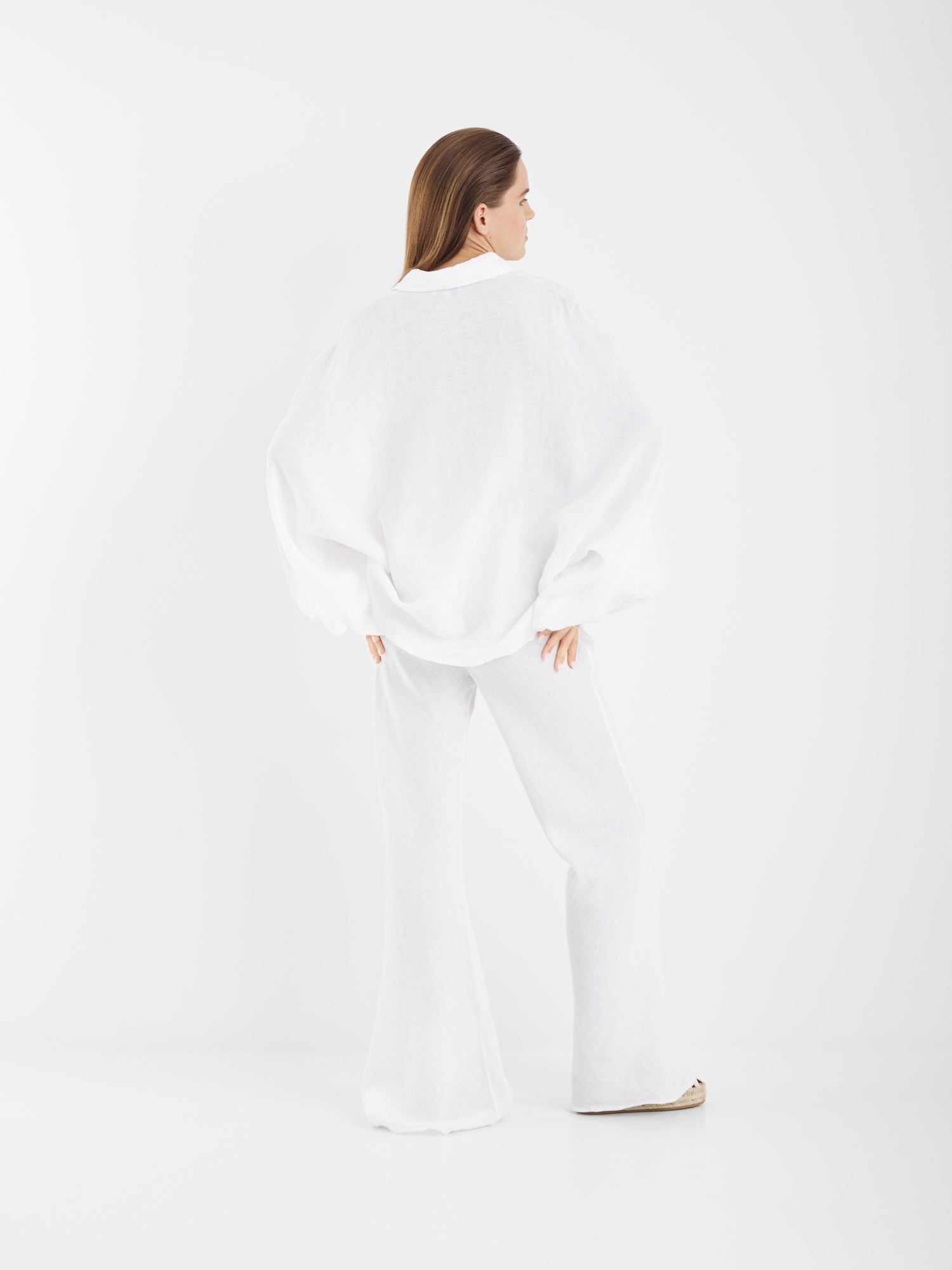 Eine Person steht mit dem Gesicht zur Wand und trägt die Bluse Antonia Linen White mit einer weißen Hose mit weitem Bein in einer minimalistischen, weißen Studioumgebung.