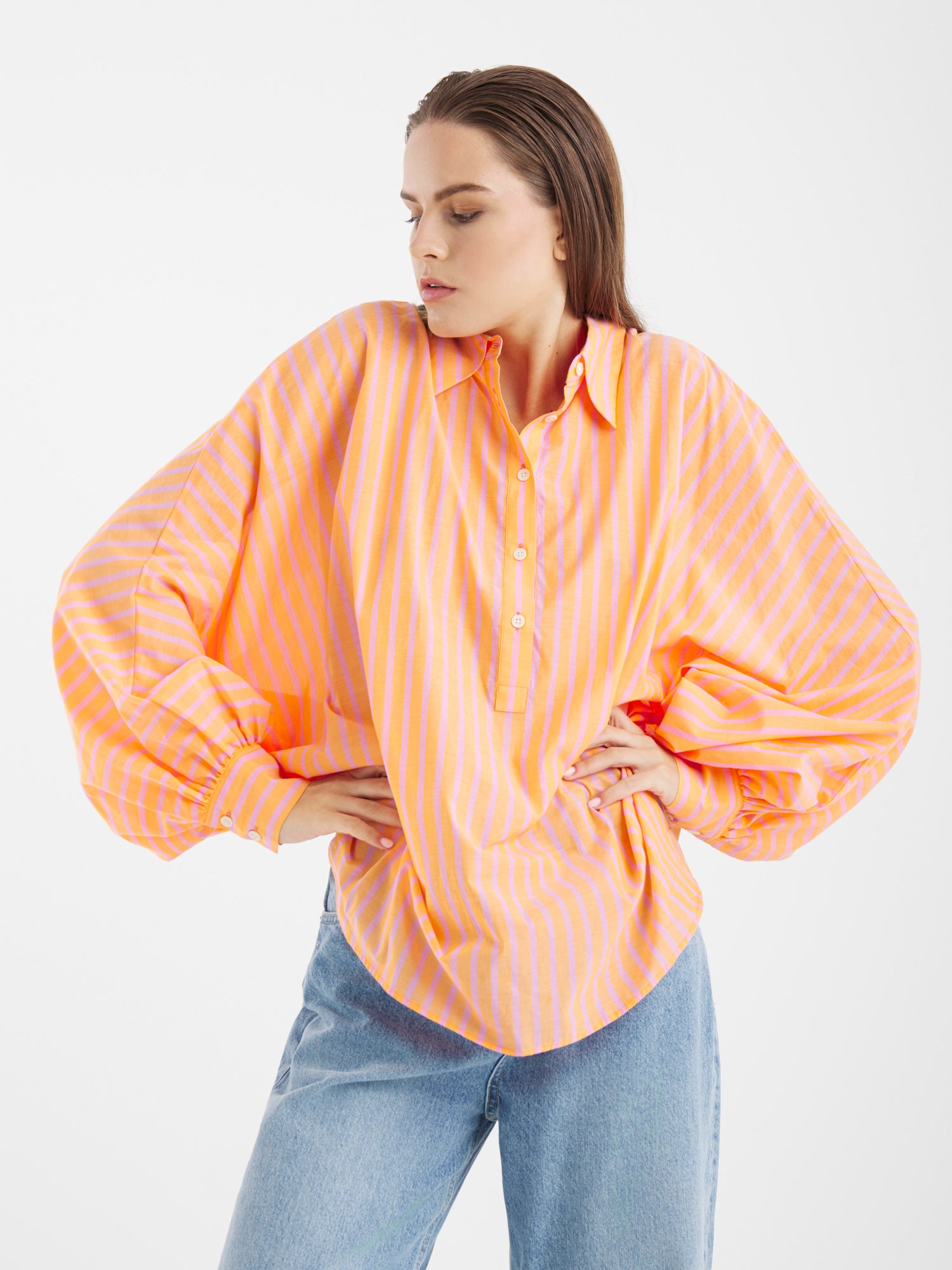 Eine Frau modelliert das Antonia Voile Coral Pink, ein locker sitzendes, langärmeliges Button-up-Hemd mit orangefarbenen und rosafarbenen Streifen, das mit einer hellblauen Jeans kombiniert wird. Sie posiert mit den Händen in den Hüften vor einem schlichten weißen Hintergrund.