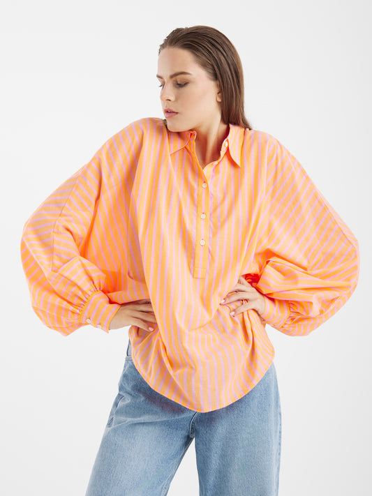 Eine Frau modelliert das Antonia Voile Coral Pink, ein locker sitzendes, langärmeliges Button-up-Hemd mit orangefarbenen und rosafarbenen Streifen, das mit einer hellblauen Jeans kombiniert wird. Sie posiert mit den Händen in den Hüften vor einem schlichten weißen Hintergrund.