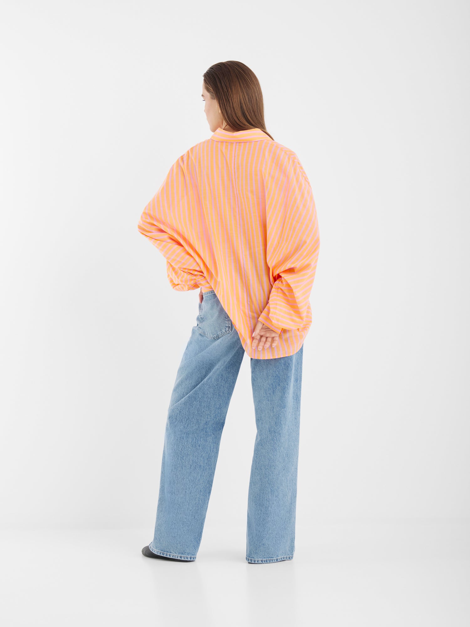 Eine Frau steht mit dem Antonia Voile Coral Pink Hemd mit weiten Ärmeln und hellblauen Jeans mit weitem Bein da. Ihr langes Haar fällt vor einem schlichten weißen Hintergrund herab.