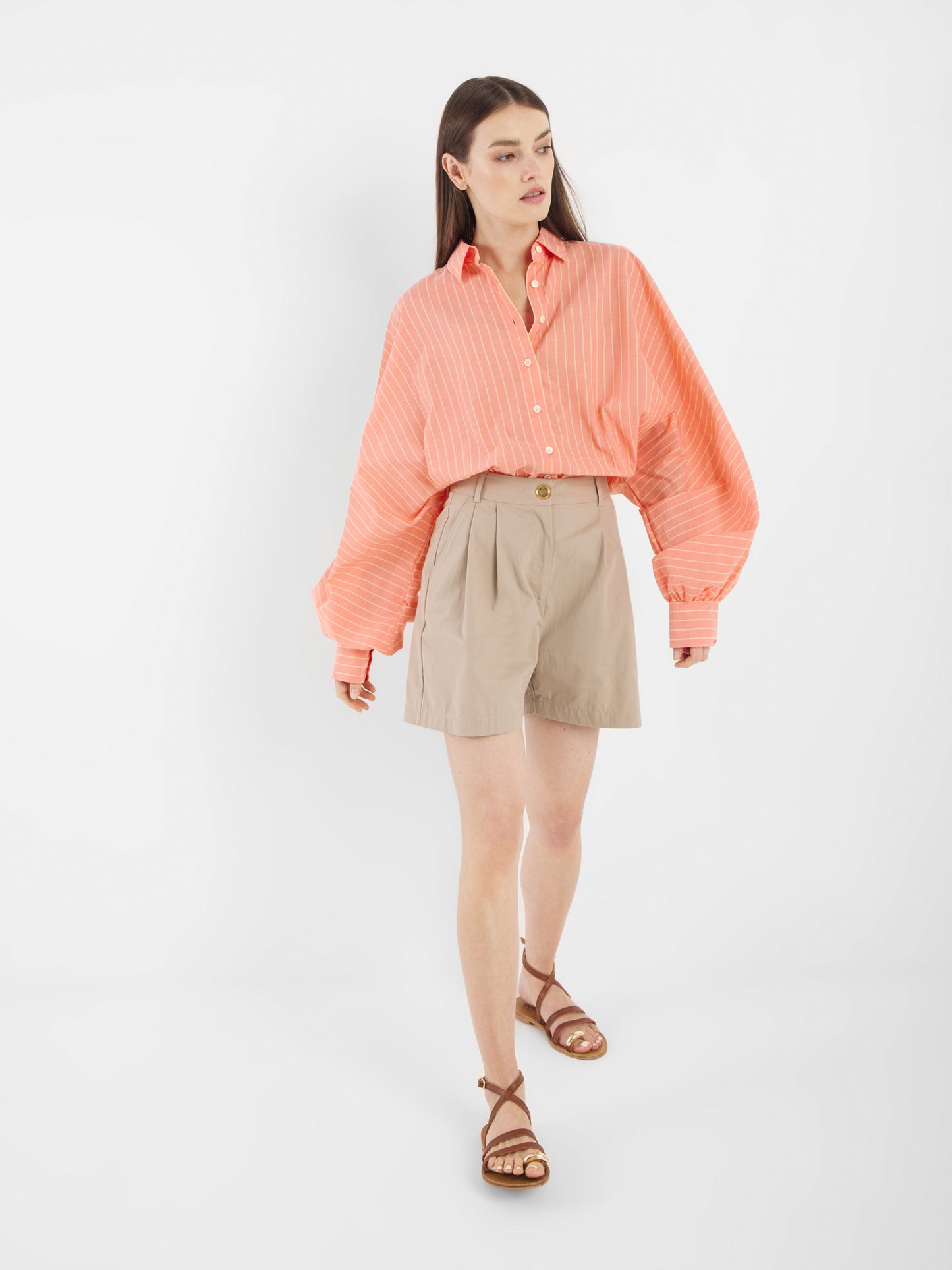 Eine Frau, die die Antonia Short Coral Stripes mit beigen Bundfaltenhosen und braunen Sandalen trägt, läuft vor einem schlichten weißen Hintergrund.