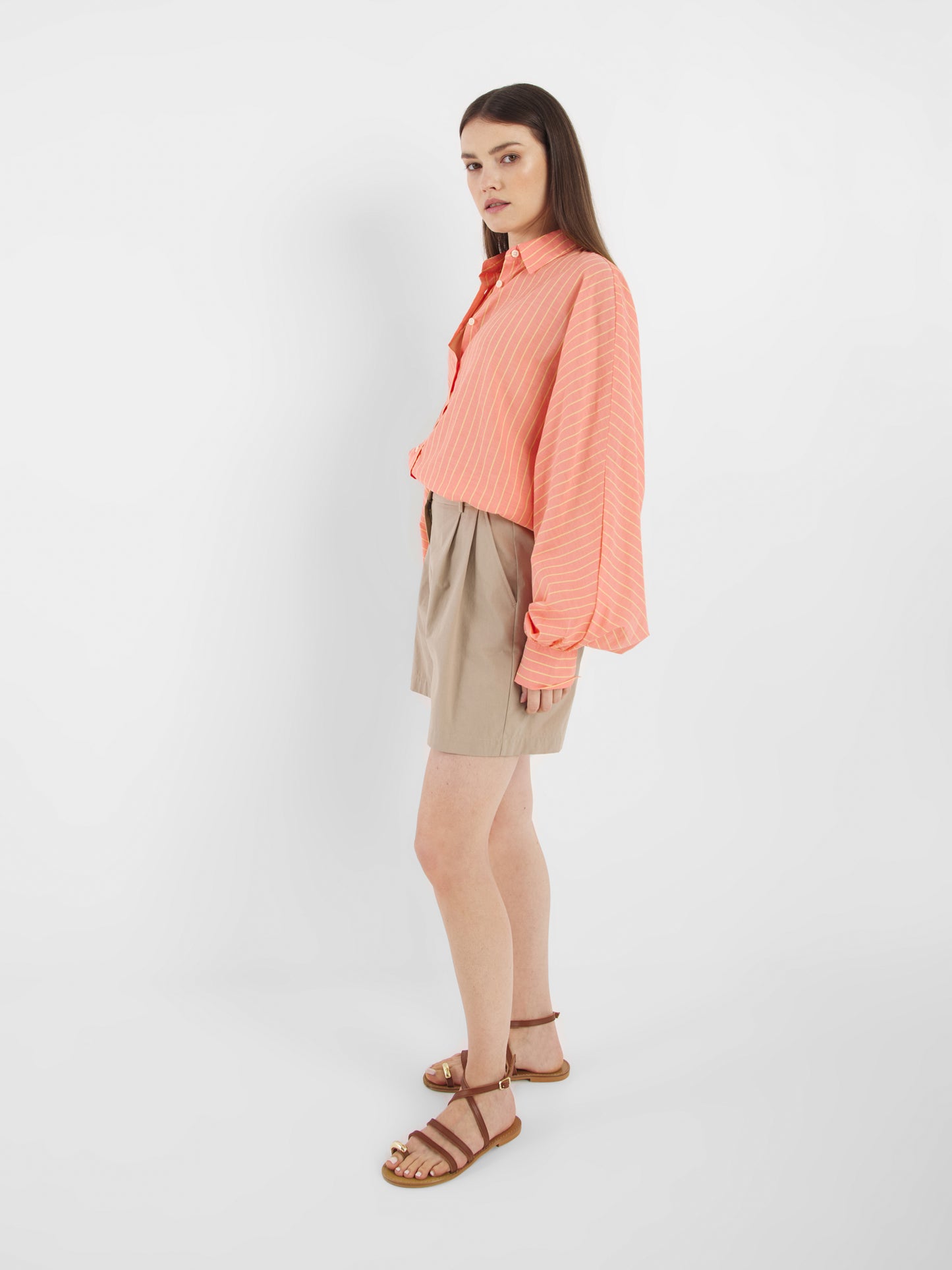 Eine Frau steht seitlich vor einem schlichten weißen Hintergrund und trägt die Antonia Short Coral Stripes mit einer lockeren korallenroten Langarmbluse und braunen Sandalen.