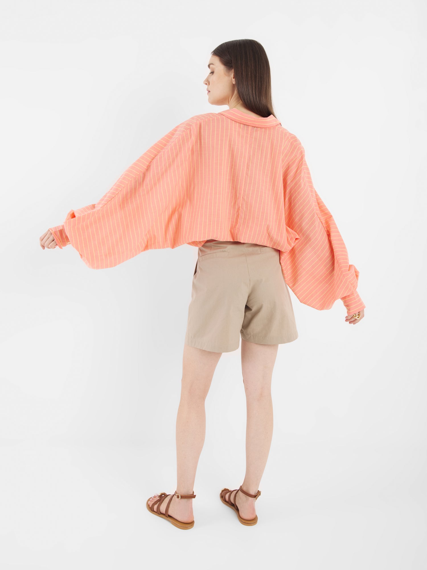 Eine Frau blickt von sich weg und trägt das Antonia Short Coral Stripes, ein lockeres pfirsichfarbenes, langärmeliges Hemd mit weißen Streifen, beigefarbene Shorts und braune Sandalen vor einem schlichten weißen Hintergrund.