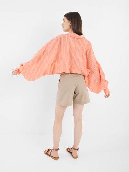 Eine Frau blickt von sich weg und trägt das Antonia Short Coral Stripes, ein lockeres pfirsichfarbenes, langärmeliges Hemd mit weißen Streifen, beigefarbene Shorts und braune Sandalen vor einem schlichten weißen Hintergrund.
