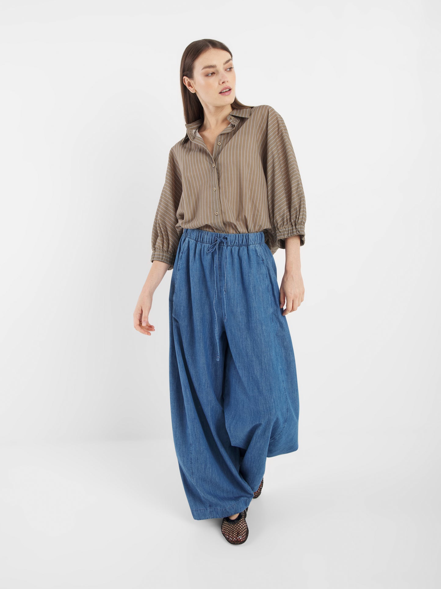 Eine Frau steht vor einem weißen Hintergrund und blickt zur Seite. Sie trägt eine blaue Lizzy-Denim-Hose mit weitem Bein und ein lockeres, braun gestreiftes Hemd, eine Hand leicht angehoben.