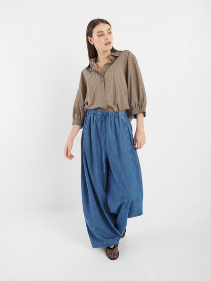 Eine Frau steht vor einem weißen Hintergrund und blickt zur Seite. Sie trägt eine blaue Lizzy-Denim-Hose mit weitem Bein und ein lockeres, braun gestreiftes Hemd, eine Hand leicht angehoben.