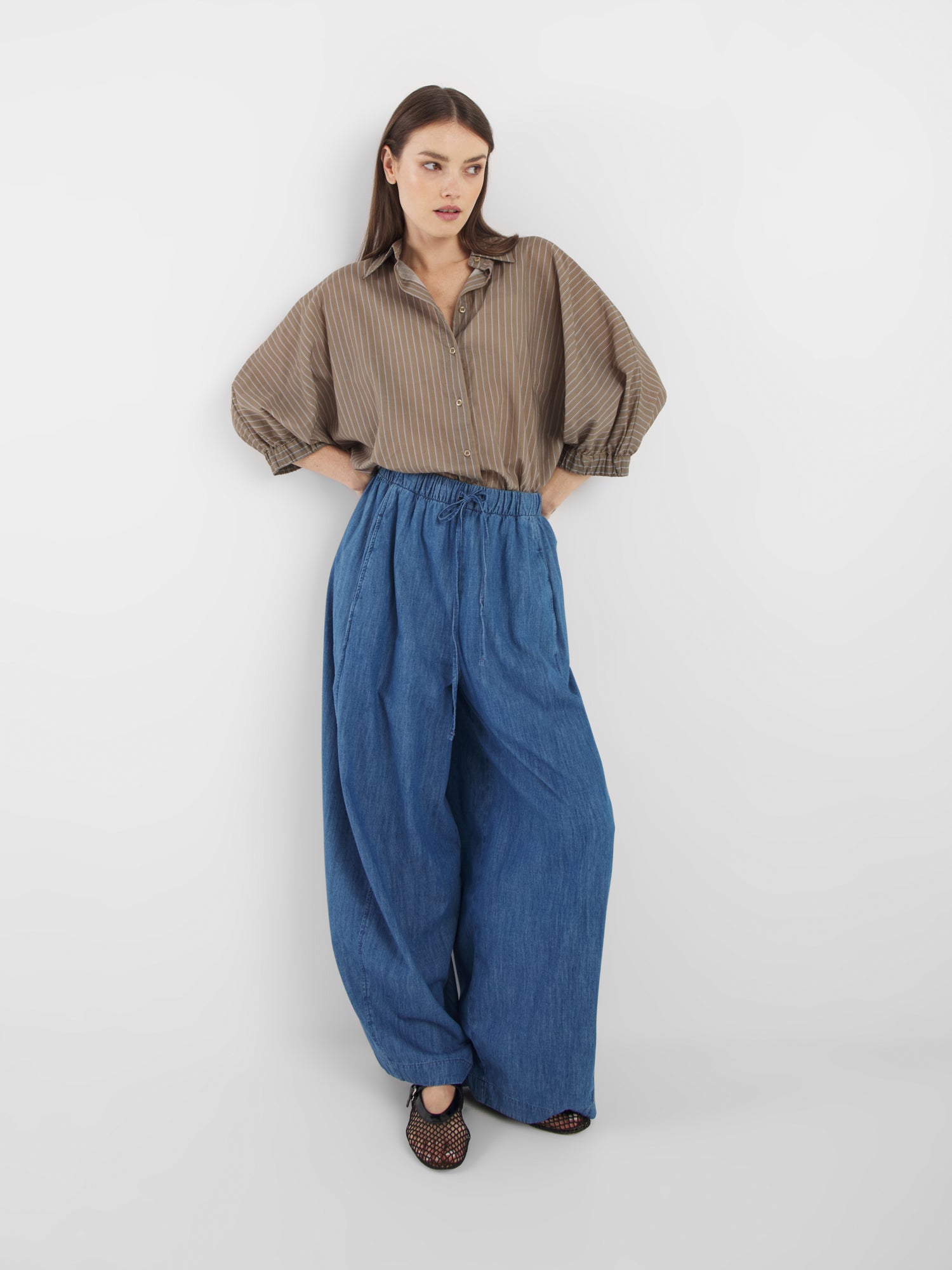 Eine Frau steht vor einem weißen Hintergrund und trägt eine locker sitzende blaue Lizzy-Denim-Hose mit weitem Bein, ein braun gestreiftes Button-up-Hemd mit voluminösen Ärmeln und schwarze gewebte Schuhe. Sie blickt leicht zur Seite und nimmt eine entspannte Haltung ein.