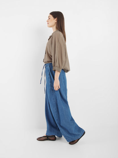 Eine Frau steht im Profil vor einem schlichten weißen Hintergrund. Sie trägt eine weite blaue Kordelzughose von Lizzy Denim, ein lockeres braun gestreiftes Hemd und schwarze Netzschuhe.