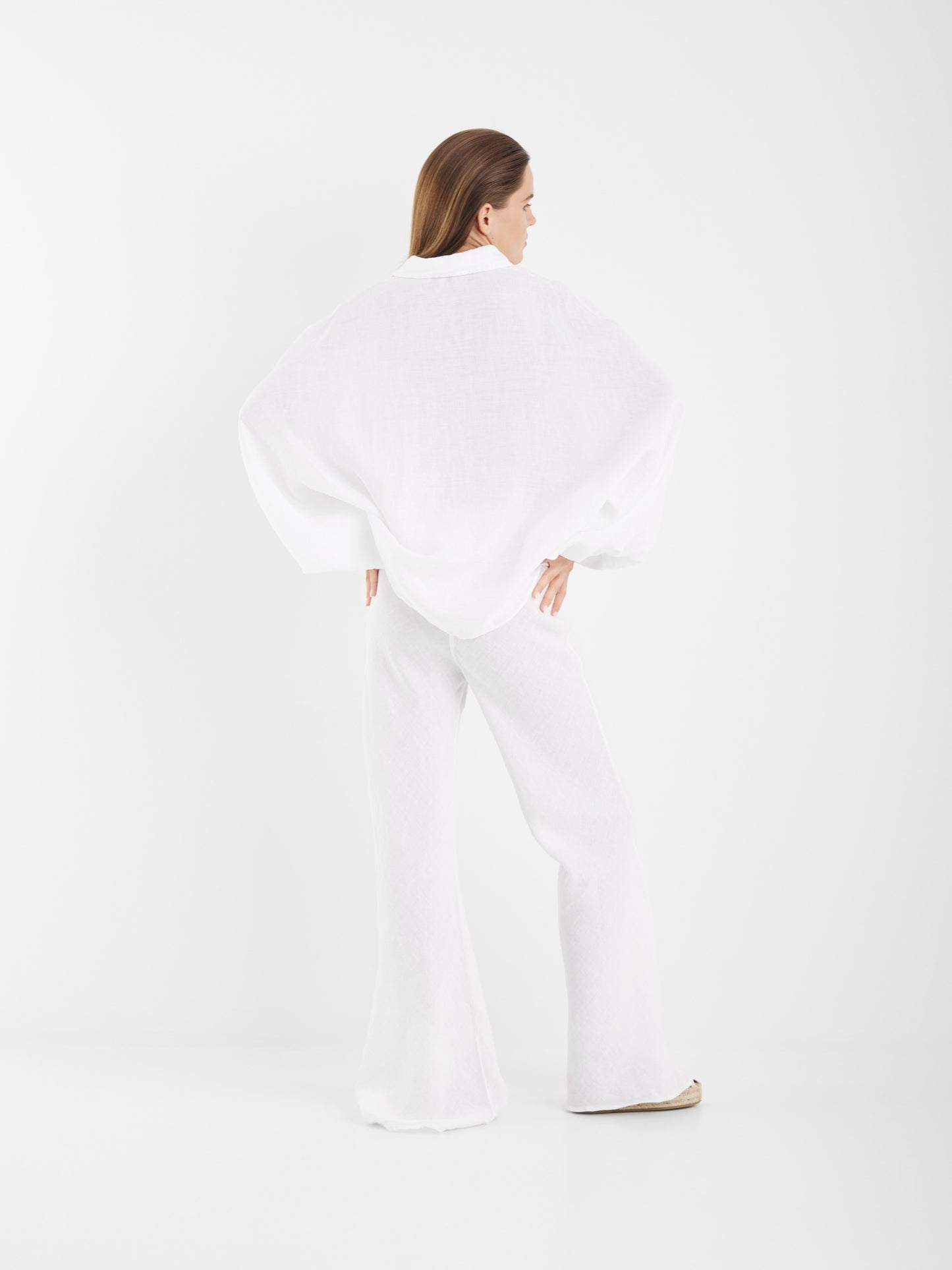 Eine Frau steht mit dem Rücken zur Kamera in einem minimalistischen weißen Studio. Sie trägt das Bluse Antonia Slim Linen White Top mit einer weißen Hose mit weitem Bein.