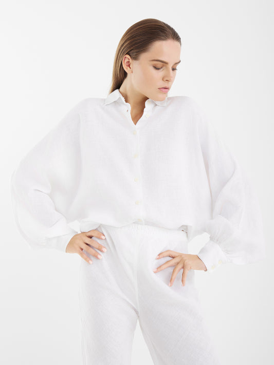 Eine Frau trägt das "White Linen ! Feels like summer", ein lockeres, langärmeliges weißes Button-up-Hemd und eine weiße Hose mit hoher Taille. Sie steht vor einem weißen Hintergrund, hat beide Hände in die Hüften gestemmt und schaut nach unten.
