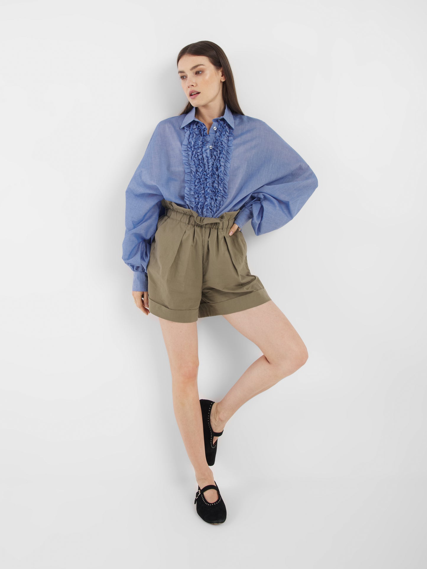 Eine Frau steht vor einem weißen Hintergrund und trägt die Antonia Slim Ruffle Denim, olivgrüne High-Waisted Shorts und schwarze Flats. Ein Bein ist leicht angewinkelt und sie stützt ihre Hand auf die Hüfte.