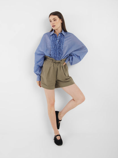 Eine Frau steht vor einem weißen Hintergrund und trägt die Antonia Slim Ruffle Denim, olivgrüne High-Waisted Shorts und schwarze Flats. Ein Bein ist leicht angewinkelt und sie stützt ihre Hand auf die Hüfte.