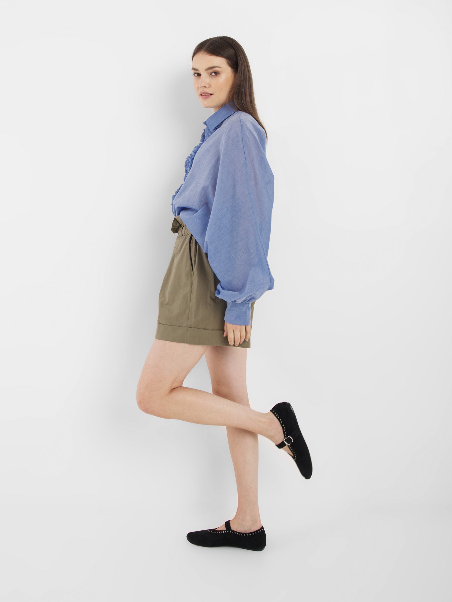 Eine Frau, die ein blaues Langarmshirt, khakifarbene Shorts von Shorts Maui und schwarze Ballerinas trägt, steht seitlich vor einem weißen Hintergrund, hebt ein Bein leicht an und blickt in die Kamera. Die Shorts von Shorts Maui sind aus hochwertigem Baumwoll-Voile gefertigt und werden in Portugal hergestellt.