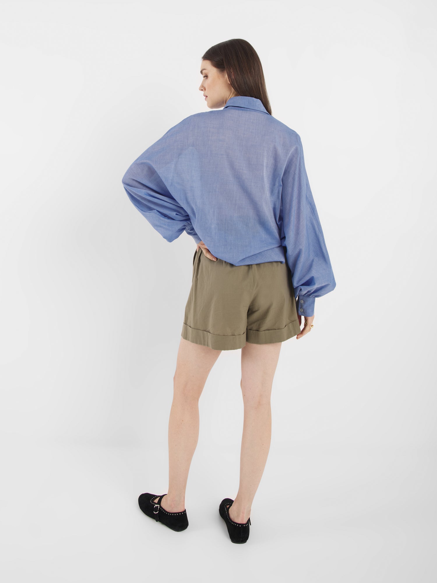 Vor einem schlichten weißen Hintergrund steht eine Frau mit dem Rücken zur Kamera; sie trägt ein lockeres blaues Baumwoll-Voile-Hemd mit Knöpfen, olivgrüne „Maui“-Shorts und schwarze Schuhe.