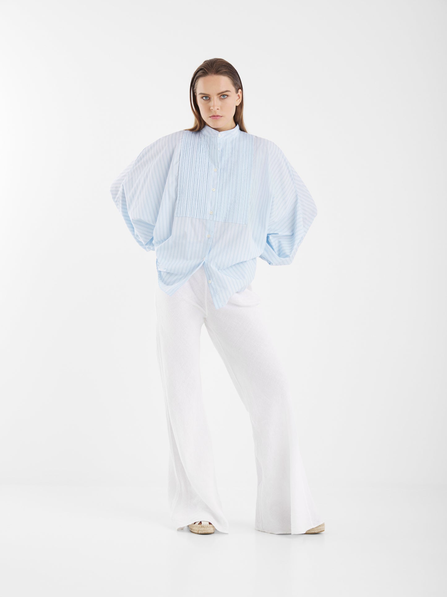 Eine Frau mit langen braunen Haaren und einem ernsten Gesichtsausdruck steht vor einem weißen Hintergrund und trägt die Antonia Smoking Blue Stripes, eine blau gestreifte Bluse in Übergröße mit weiten Ärmeln im Smoking-Stil, kombiniert mit einer weißen Hose mit weitem Bein.