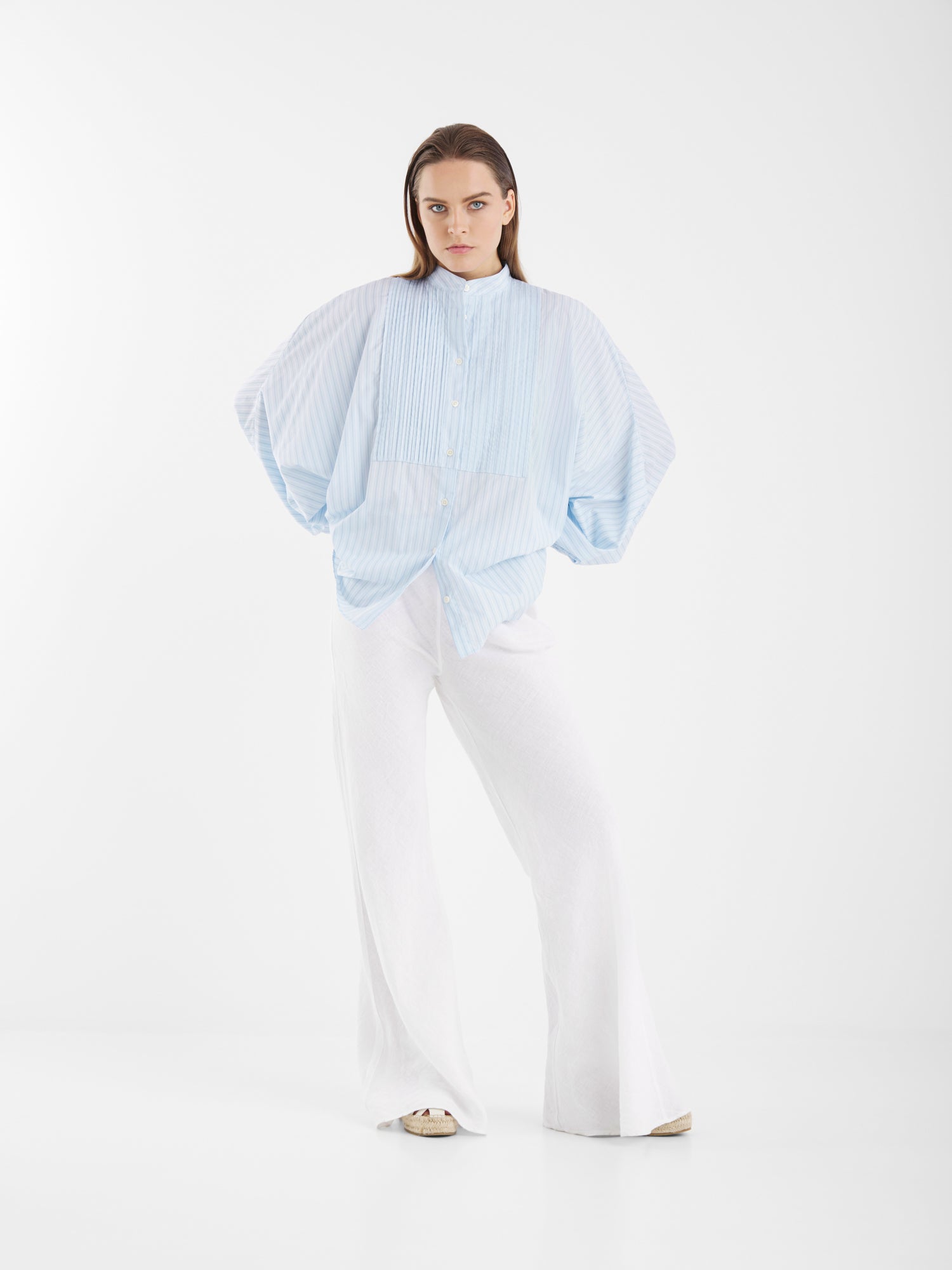 Eine Frau mit langen braunen Haaren und einem ernsten Gesichtsausdruck steht vor einem weißen Hintergrund und trägt die Antonia Smoking Blue Stripes, eine blau gestreifte Bluse in Übergröße mit weiten Ärmeln im Smoking-Stil, kombiniert mit einer weißen Hose mit weitem Bein.