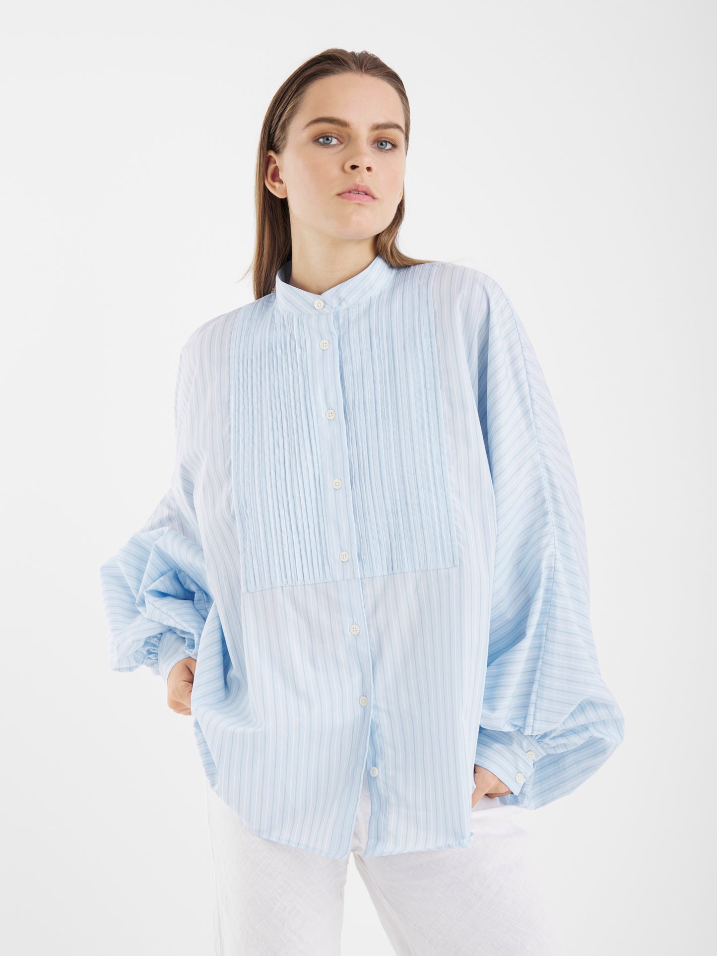 Eine Person trägt die Antonia Smoking Blue Stripes Bluse - ein übergroßes Damenhemd mit hellblauen und weißen Streifen sowie Smokingfalten - zusammen mit einer weißen Hose vor einem schlichten weißen Hintergrund.