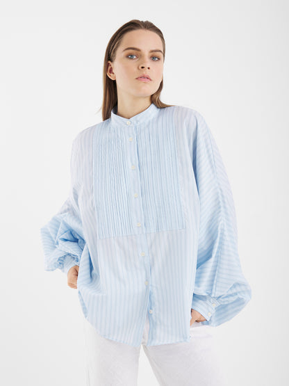 Eine Person trägt die Antonia Smoking Blue Stripes Bluse - ein übergroßes Damenhemd mit hellblauen und weißen Streifen sowie Smokingfalten - zusammen mit einer weißen Hose vor einem schlichten weißen Hintergrund.