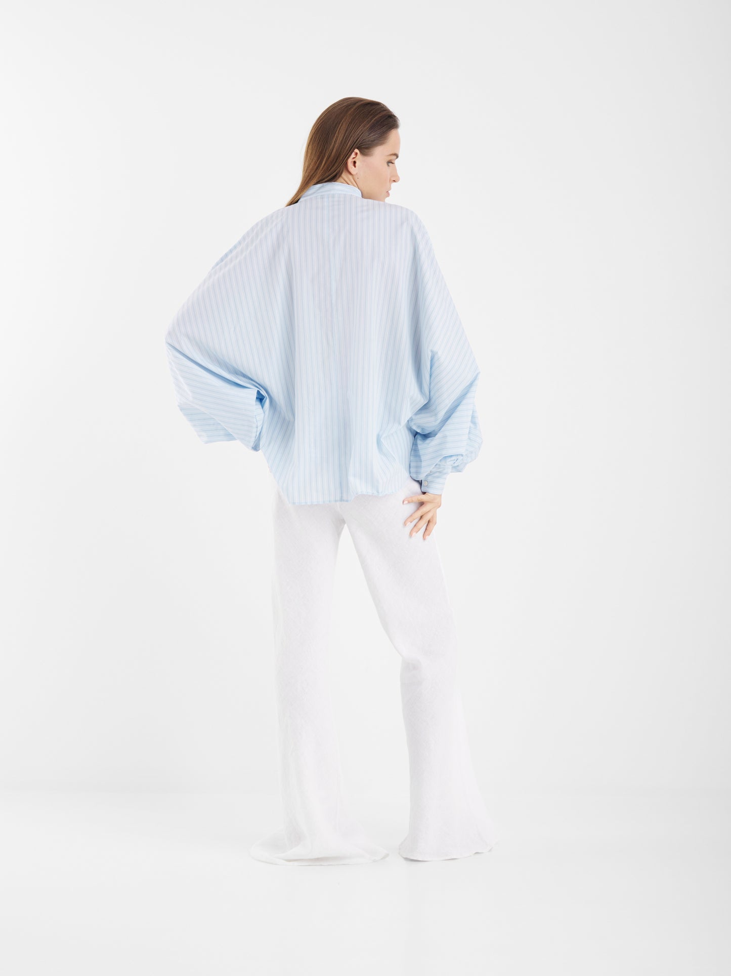 Eine Frau steht mit dem Rücken zur Kamera und trägt die Antonia Smoking Blue Stripes Bluse und weiße Schlaghosen, die vor einem schlichten weißen Hintergrund einen schicken Oversized-Look ergeben.