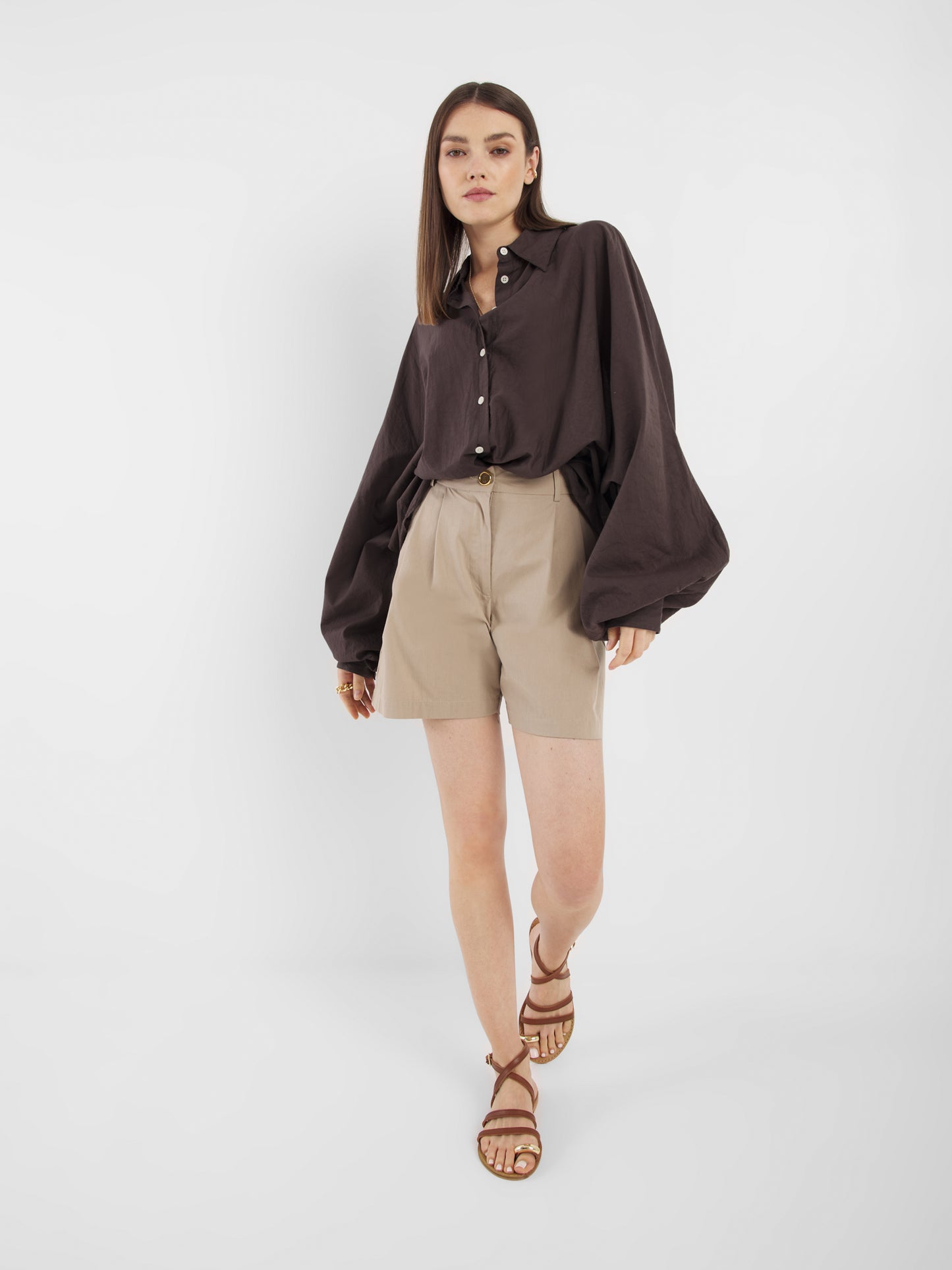 Eine Frau mit glattem Haar und neutralem Gesichtsausdruck steht vor einem weißen Hintergrund und trägt das Antonia Voile Brown, ein lockeres dunkelbraunes Button-up-Hemd mit Ballonärmeln, gepaart mit beigen High-Waisted-Shorts und braunen Riemchensandalen.