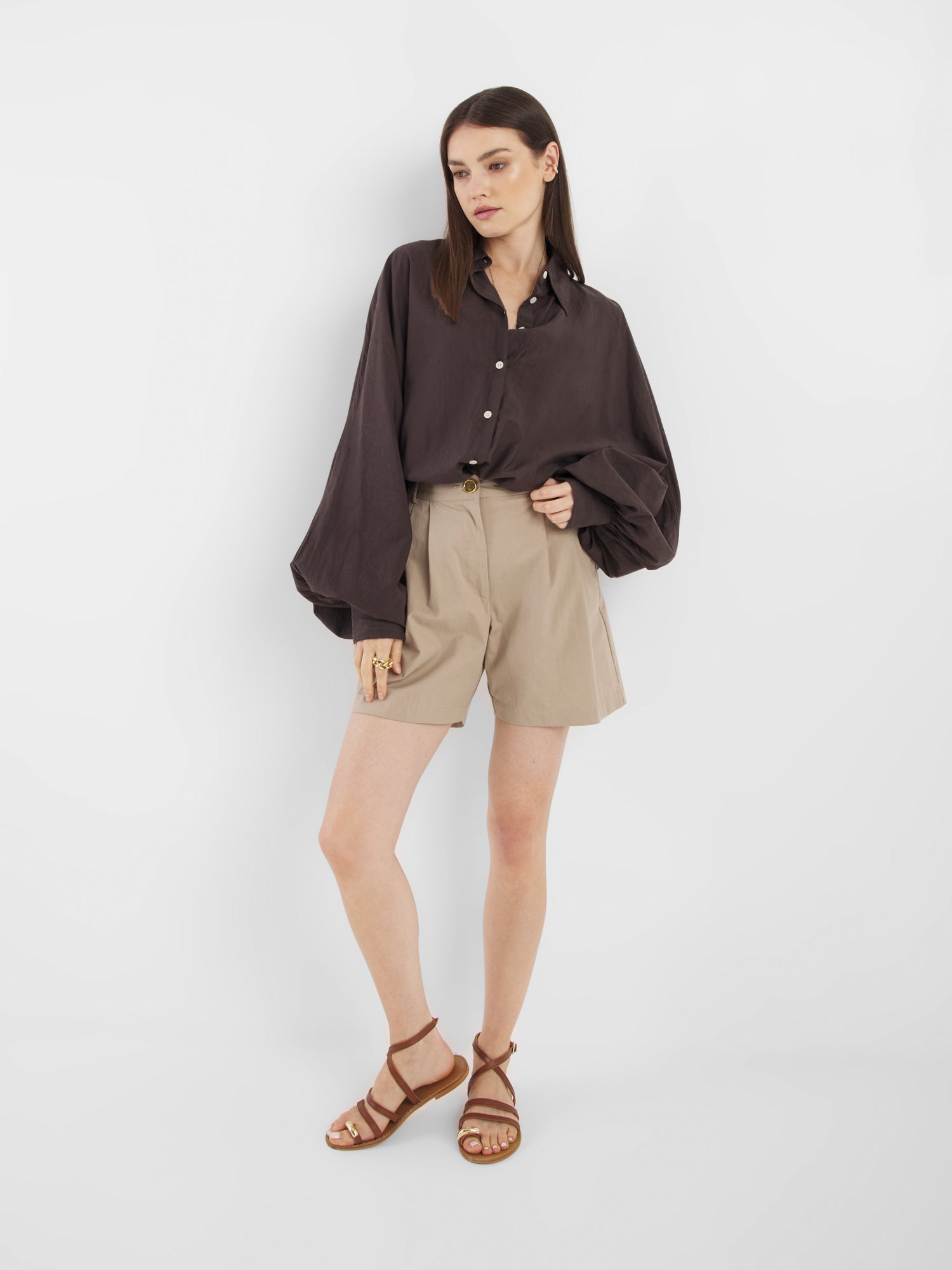 Eine Frau mit glattem braunem Haar und neutralem Gesichtsausdruck modelliert Shorts Safari - beigefarbene Shorts mit hoher Taille - in Kombination mit einem lockeren, dunkelbraunen Button-up-Shirt und braunen Riemchensandalen vor einem weißen Hintergrund.
