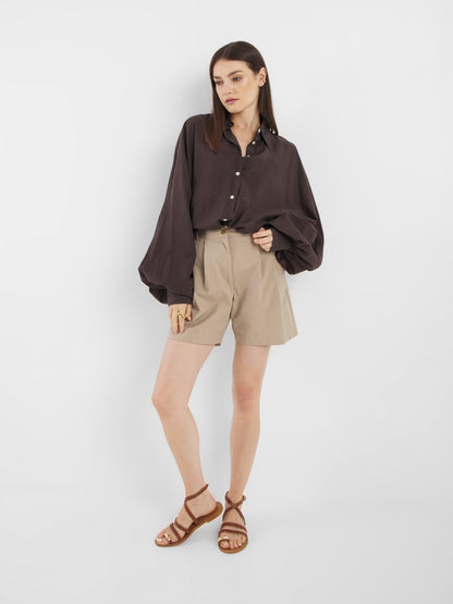 Eine Frau mit glattem braunem Haar und neutralem Gesichtsausdruck modelliert Shorts Safari - beigefarbene Shorts mit hoher Taille - in Kombination mit einem lockeren, dunkelbraunen Button-up-Shirt und braunen Riemchensandalen vor einem weißen Hintergrund.