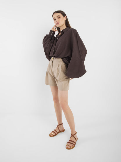 Eine Frau posiert vor einem weißen Hintergrund in der Antonia Voile Brown, einem lockeren dunkelbraunen Button-up-Hemd, gepaart mit beigen, hoch taillierten Shorts und braunen Riemchensandalen. Sie hat glattes Haar und eine Hand neben ihrem Gesicht.