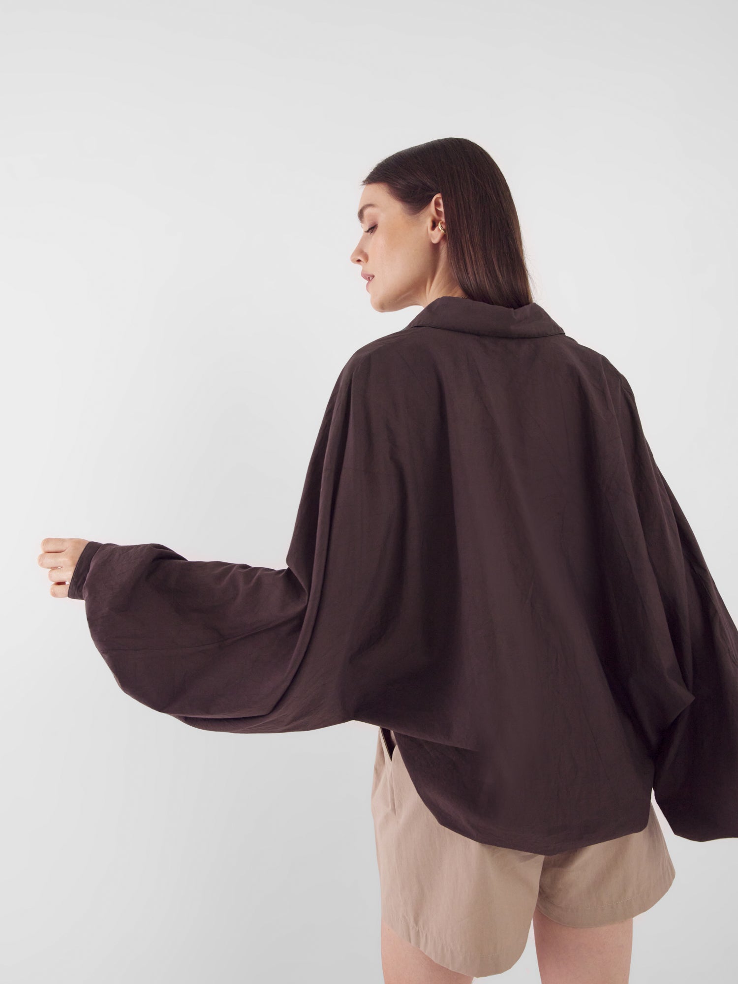 Eine Frau steht seitlich vor einem schlichten Hintergrund. Sie trägt das lockere, langärmelige Hemd Antonia Voile Brown mit weiten Ärmeln und hellbeigefarbene Shorts. Ihr Kopf ist leicht gedreht und ihr rechter Arm ist ausgestreckt.