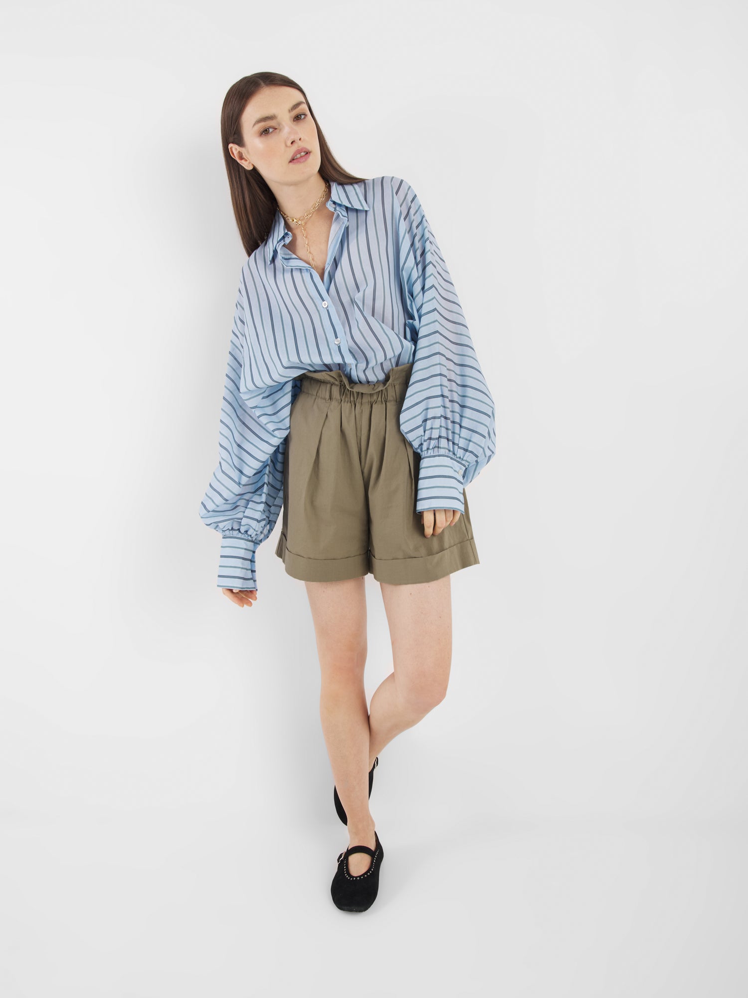 Eine Frau mit glattem braunem Haar und einer entspannten Pose steht vor einem weißen Hintergrund und trägt die Bluse Antonia Voile Blue Stripes, khakifarbene Shorts mit hoher Taille, schwarze Schuhe und eine Halskette.