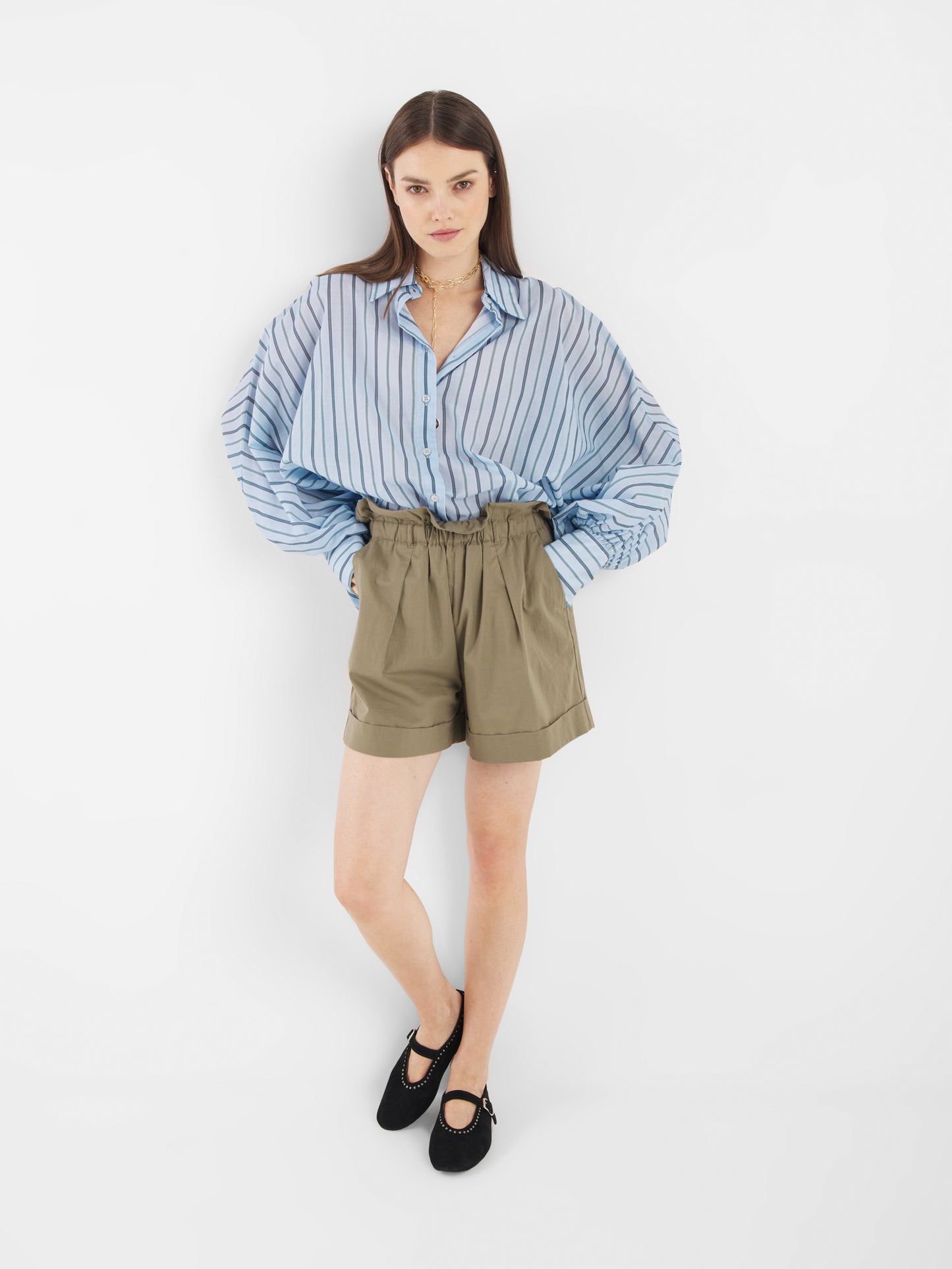 Eine junge Frau steht vor einem weißen Hintergrund, trägt die Bluse Antonia Voile Blue Stripes, khakifarbene, hochtaillierte Papiertaschenshorts und schwarze Mary-Jane-Schuhe und schaut mit neutralem Blick in die Kamera.