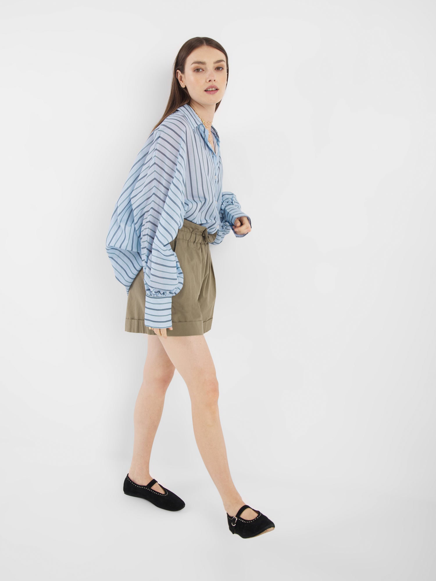 Eine Frau mit langen braunen Haaren trägt die Bluse Antonia Voile Blue Stripes in Übergröße, die sie in eine hoch taillierte Khaki-Shorts mit schwarzen Schuhen gesteckt hat. Sie geht vor einem schlichten weißen Hintergrund nach vorne.