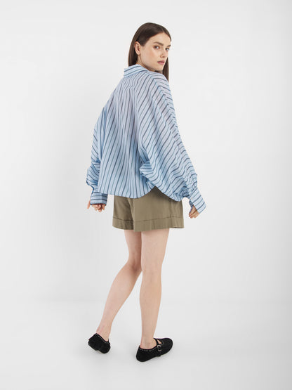 Eine Frau mit langen braunen Haaren trägt die Antonia Voile Blue Stripes Bluse und khakifarbene Shorts. Sie steht leicht gedreht und blickt über ihre Schulter, gepaart mit schwarzen Schuhen vor einem schlichten weißen Hintergrund.